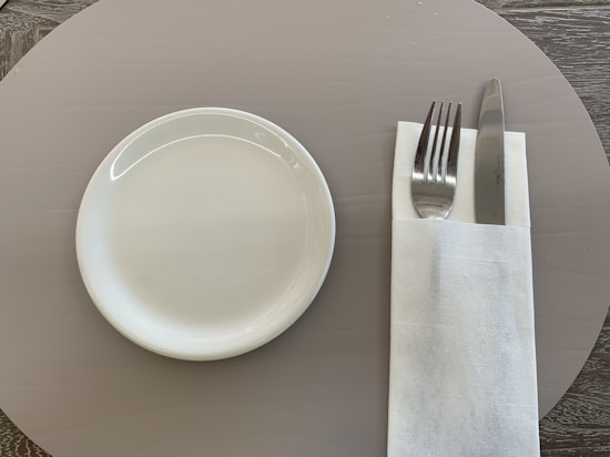 A neatly arranged place setting on a table includes a small white plate on the left and a folded white napkin on the right, which holds a fork and a knife beside each other.