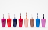 Eight nail polish bottles in various bright colors are arranged in a row against a plain white background. The bottles feature shades including pink, red, dark blue, light orange, brown, light blue, and purple. Each bottle has a visible brush sticking out, showcasing the precise design typical for nail polish containers.