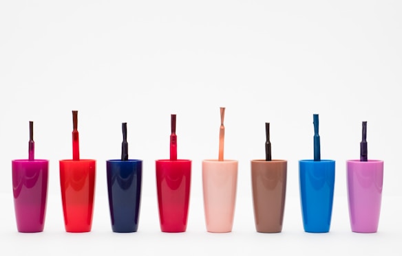 Eight nail polish bottles in various bright colors are arranged in a row against a plain white background. The bottles feature shades including pink, red, dark blue, light orange, brown, light blue, and purple. Each bottle has a visible brush sticking out, showcasing the precise design typical for nail polish containers.