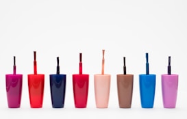 Eight nail polish bottles in various bright colors are arranged in a row against a plain white background. The bottles feature shades including pink, red, dark blue, light orange, brown, light blue, and purple. Each bottle has a visible brush sticking out, showcasing the precise design typical for nail polish containers.