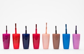 Eight nail polish bottles in various bright colors are arranged in a row against a plain white background. The bottles feature shades including pink, red, dark blue, light orange, brown, light blue, and purple. Each bottle has a visible brush sticking out, showcasing the precise design typical for nail polish containers.