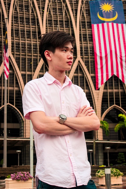 A person standing with arms crossed in front of a building with intricate architectural details. Two flags with crescent moons and stars are visible, indicating a Malaysian context. The person is wearing a light pink shirt and a wristwatch, looking towards the side.