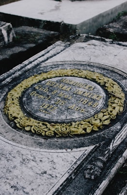 A weathered, ornate tombstone with detailed carvings and engraved inscriptions. It features an elliptical design bordered by a wreath-like arrangement of yellow tinted floral carvings. The stone has a worn, aged appearance, suggesting it has been exposed to the elements over time.