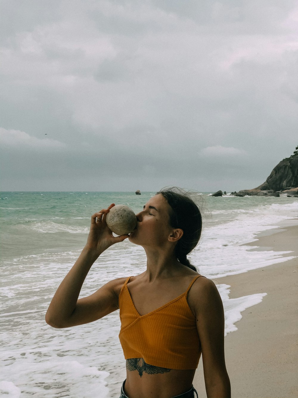 Mujer en camiseta sin mangas naranja sosteniendo una piedra gris cerca del mar durante el día