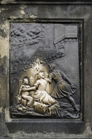 An intricately carved relief depicting a historical scene with a knight engaging with a woman and child. The background features a medieval castle with people on the battlements, possibly amidst a siege or battle. The artwork is set in a stone frame, with detailed expressions and textures.