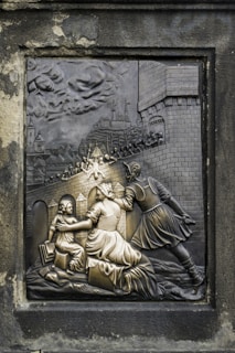 An intricately carved relief depicting a historical scene with a knight engaging with a woman and child. The background features a medieval castle with people on the battlements, possibly amidst a siege or battle. The artwork is set in a stone frame, with detailed expressions and textures.