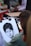 A person is sketching a detailed portrait with charcoal or pencil on a drawing pad. The portrait is of a person with short dark hair and a calm expression. Various art supplies and other people are visible in the background, suggesting an outdoor or public setting.