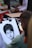 A person is sketching a detailed portrait with charcoal or pencil on a drawing pad. The portrait is of a person with short dark hair and a calm expression. Various art supplies and other people are visible in the background, suggesting an outdoor or public setting.