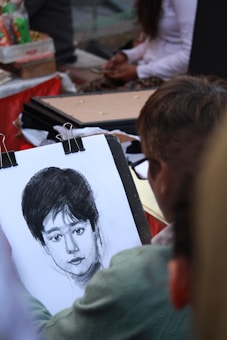 A person is sketching a detailed portrait with charcoal or pencil on a drawing pad. The portrait is of a person with short dark hair and a calm expression. Various art supplies and other people are visible in the background, suggesting an outdoor or public setting.