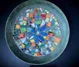 A circular mosaic artwork featuring an intricate design of various colorful tiles arranged on a metallic platter. The center is dominated by a blue flower-like pattern surrounded by small, colorful square and rectangular tiles in shades of orange, green, purple, and red. Each tile is carefully placed to form a harmonious and vibrant composition.