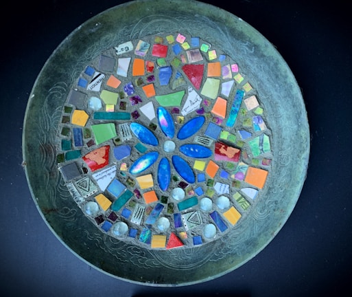 A circular mosaic artwork featuring an intricate design of various colorful tiles arranged on a metallic platter. The center is dominated by a blue flower-like pattern surrounded by small, colorful square and rectangular tiles in shades of orange, green, purple, and red. Each tile is carefully placed to form a harmonious and vibrant composition.