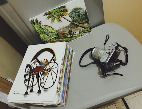 A table holds a pile of paintings and a camera. One painting features a horse-drawn carriage, while another depicts a lush landscape with trees and a person near a river. The camera is silver with a black strap and has a vintage design.