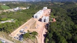 Aerial view of a construction site surrounded by a dense forest. Several large, multi-story concrete buildings are in progress, with visible construction materials and equipment scattered around. The surrounding landscape includes green fields, trees, and distant hills.