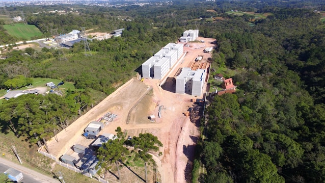 Aerial view of a construction site surrounded by a dense forest. Several large, multi-story concrete buildings are in progress, with visible construction materials and equipment scattered around. The surrounding landscape includes green fields, trees, and distant hills.
