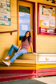 A person sitting casually on a yellow and red wooden platform outside a building. The person is wearing a red and white striped shirt, blue jeans, and white sneakers. Behind is a glass door with a sign for the Downtown Issaquah Association and a community bulletin board with various colorful flyers and notices.
