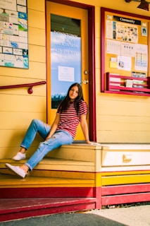 A person sitting casually on a yellow and red wooden platform outside a building. The person is wearing a red and white striped shirt, blue jeans, and white sneakers. Behind is a glass door with a sign for the Downtown Issaquah Association and a community bulletin board with various colorful flyers and notices.
