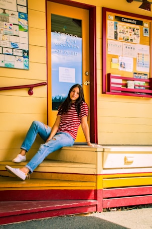 A person sitting casually on a yellow and red wooden platform outside a building. The person is wearing a red and white striped shirt, blue jeans, and white sneakers. Behind is a glass door with a sign for the Downtown Issaquah Association and a community bulletin board with various colorful flyers and notices.