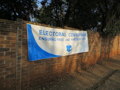 A large white and blue banner is stretched across a brick wall. The banner reads 'ELECTORAL COMMISSION ENSURING FREE AND FAIR ELECTIONS' with the logo of the Independent Electoral Commission of South Africa in the center. The surrounding area is shaded by trees.