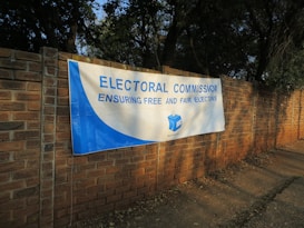 A large white and blue banner is stretched across a brick wall. The banner reads 'ELECTORAL COMMISSION ENSURING FREE AND FAIR ELECTIONS' with the logo of the Independent Electoral Commission of South Africa in the center. The surrounding area is shaded by trees.