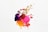 A splash of colorful paints, including purple, pink, gold, red, and yellow, creates an abstract design against a white background. The paint splatters and drips are randomly distributed, forming an energetic and dynamic pattern.