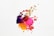 A splash of colorful paints, including purple, pink, gold, red, and yellow, creates an abstract design against a white background. The paint splatters and drips are randomly distributed, forming an energetic and dynamic pattern.