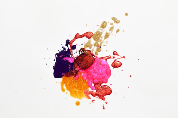 A splash of colorful paints, including purple, pink, gold, red, and yellow, creates an abstract design against a white background. The paint splatters and drips are randomly distributed, forming an energetic and dynamic pattern.
