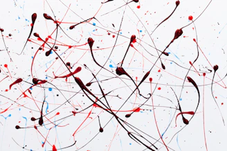 Abstract art composed of dynamic splatters and drips of red, black, and blue paint against a white background. The paint is applied in an energetic, chaotic manner with intersecting lines and scattered dots.