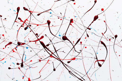 Abstract art composed of dynamic splatters and drips of red, black, and blue paint against a white background. The paint is applied in an energetic, chaotic manner with intersecting lines and scattered dots.