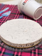 A round dessert with a light, creamy texture and speckled with small dark bits, likely a type of cheesecake or pie, sits on a glass plate. The background features a red and black checkered cloth and a tipped-over white container with ingredients.