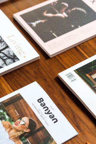 Several lifestyle magazines are arranged on a wooden table. The publications have vibrant covers, including one with the title 'Banyan' and an image of a woman wearing sunglasses and stylish earrings. The photographed magazines are placed in a neat, overlapping pattern.