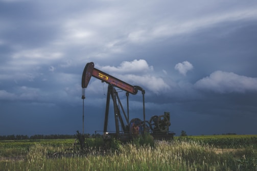 An oil pump jack is situated in a field of tall grass under a cloudy sky. The machinery is rusted and weathered, highlighting its age and frequent use. The horizon is filled with thick, dark clouds, suggesting an incoming storm. The foreground features a blend of green vegetation and field grasses swaying in the wind.