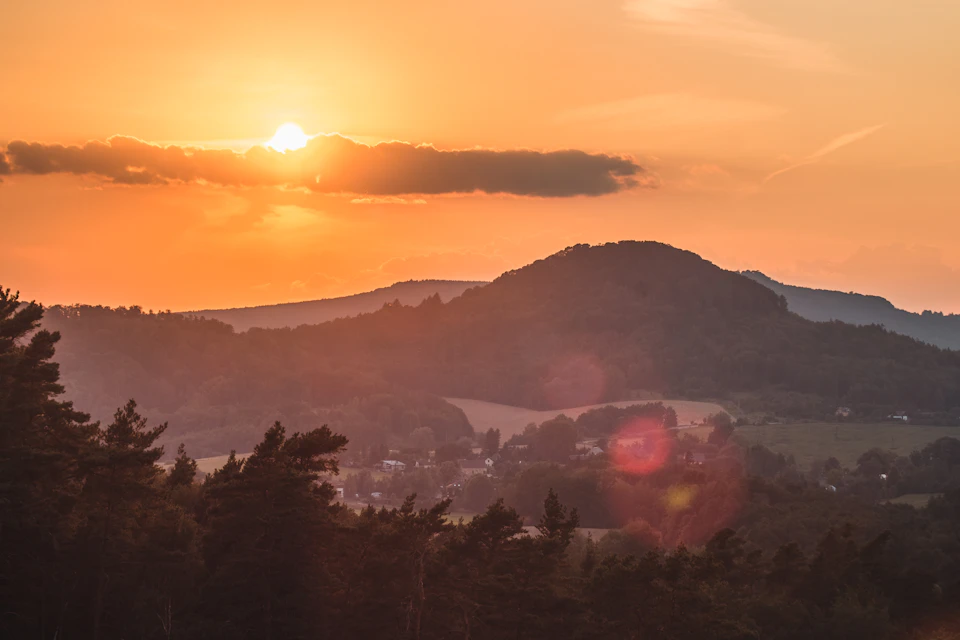 The image depicts a breathtaking sunset over a verdant hillside, with the sun setting behind a mountain range and casting a warm glow across the landscape. The sky is painted in hues of orange and yellow, creating a stunning backdrop for the scene. The hillside is lush with trees and vegetation, adding depth to the image. In the distance, houses can be seen nestled among the greenery, blending harmoniously with nature's palette.

The perspective of the photo is from a high vantage point, providing an expansive view of the landscape below. The sun appears to be setting behind the mountain range, casting long shadows and bathing everything in its warm light.