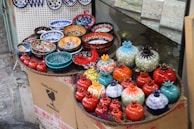 A display of colorful ceramic bowls and decorative items resembling pomegranates are arranged on a raised surface. The objects feature intricate patterns and vibrant colors, including red, blue, orange, and turquoise. They are placed on top of cardboard packaging with labels indicating fragility.