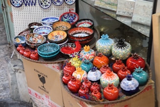 A display of colorful ceramic bowls and decorative items resembling pomegranates are arranged on a raised surface. The objects feature intricate patterns and vibrant colors, including red, blue, orange, and turquoise. They are placed on top of cardboard packaging with labels indicating fragility.