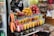 A vibrant fruit juice stand displays an array of freshly prepared juices in clear plastic cups, stacked neatly in rows. Bright oranges, pomegranates, and other fruits are piled on a metal tray above the juices. Whole carrots, along with containers filled with sliced watermelon, papaya, and other fruits, can be seen. Various packaged snacks are displayed in the background, adding to the assortment available for sale.