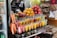 A vibrant fruit juice stand displays an array of freshly prepared juices in clear plastic cups, stacked neatly in rows. Bright oranges, pomegranates, and other fruits are piled on a metal tray above the juices. Whole carrots, along with containers filled with sliced watermelon, papaya, and other fruits, can be seen. Various packaged snacks are displayed in the background, adding to the assortment available for sale.