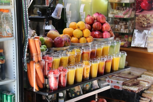 A vibrant fruit juice stand displays an array of freshly prepared juices in clear plastic cups, stacked neatly in rows. Bright oranges, pomegranates, and other fruits are piled on a metal tray above the juices. Whole carrots, along with containers filled with sliced watermelon, papaya, and other fruits, can be seen. Various packaged snacks are displayed in the background, adding to the assortment available for sale.