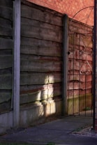 Sunlight casts intricate shadows of a wrought iron gate onto a wooden fence. The scene includes a weathered wooden fence, a pathway, and an ornate metal gate that partially conceals the view beyond.