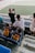 Several spectators sit in a stadium overlooking a large soccer field. Two children are wearing jerseys with the name and number 'Messi 10' on them. A man with a shaved head stands near a railing taking photos or videos with a smartphone. The seating area consists of blue chairs. Another person stands in a floral dress nearby. The atmosphere is casual and relaxed as people enjoy the view.
