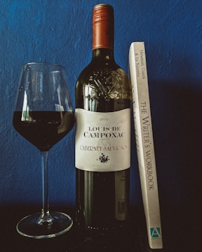 A bottle of Louis de Camponac Cabernet Sauvignon stands next to a filled wine glass, with a book titled 'The Writer's Workbook' propped up beside it, set against a textured blue wall backdrop.