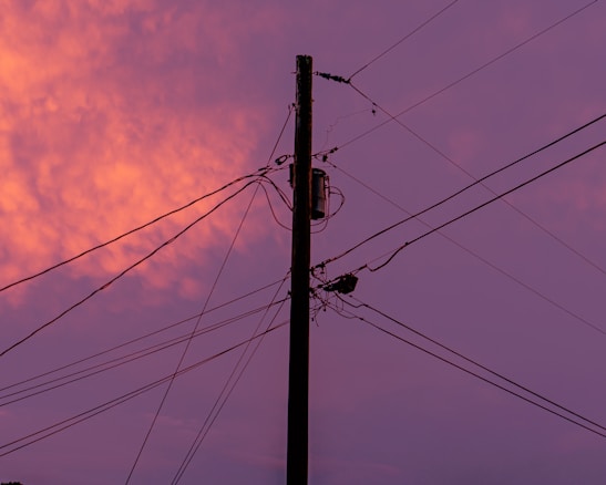 An electric pole with multiple power lines set against a vibrant sunset sky, blending shades of orange and purple hues.