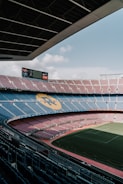 A vast football stadium with rows of seats in red and blue, and a large field in the center. The structure is open-air with a clear sky visible. Large screens are mounted at one end of the seating area, and the seats display a pattern forming a notable emblem.