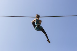 A person climbing or hanging from a tightrope, wearing a teal tank top and black leggings, against a clear blue sky.