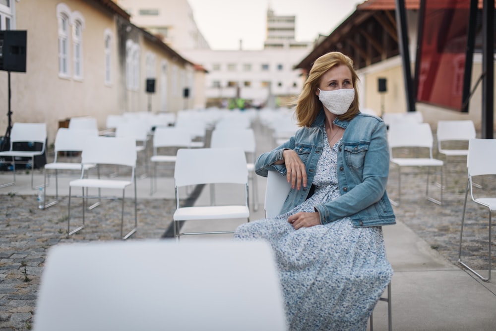 Frau in blauer Jeansjacke und weiß-schwarzem Kleid sitzt auf weißem Betonzaun während