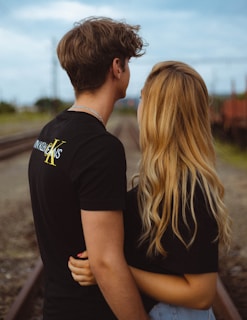 A couple stands closely on a railroad track, embracing each other. The individual on the left has short brown hair and wears a black shirt with text. The person on the right has long blonde hair and is also wearing a black top. The background features railway tracks and blurred greenery under a cloudy sky.