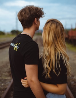 A couple stands closely on a railroad track, embracing each other. The individual on the left has short brown hair and wears a black shirt with text. The person on the right has long blonde hair and is also wearing a black top. The background features railway tracks and blurred greenery under a cloudy sky.