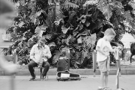 A street musician plays a saxophone while sitting on a chair in front of large tropical plants. A boy in the foreground is blurred and appears to be playing on a scooter. There is an open saxophone case and a speaker next to the musician.