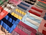A colorful patchwork quilt made up of various fabric strips sewn together, featuring a variety of patterns and textures. The fabrics display a mix of geometric and floral prints, including checks, stripes, and paisley designs.