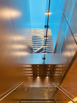 A modern interior space featuring a glass staircase leading upwards with stainless steel railings. Above, a reflection of a contemporary building with multiple floors is visible against a bright blue sky. Recessed ceiling lights illuminate the space, creating warm reflections on the metallic and glass surfaces.