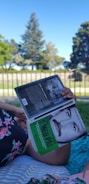 A person is lying on a blanket on a grassy area, reading a book titled 'O Meu Pai' with a photograph of a man on the cover. Surrounding the scene are trees and a clear blue sky, suggesting a peaceful outdoor setting.